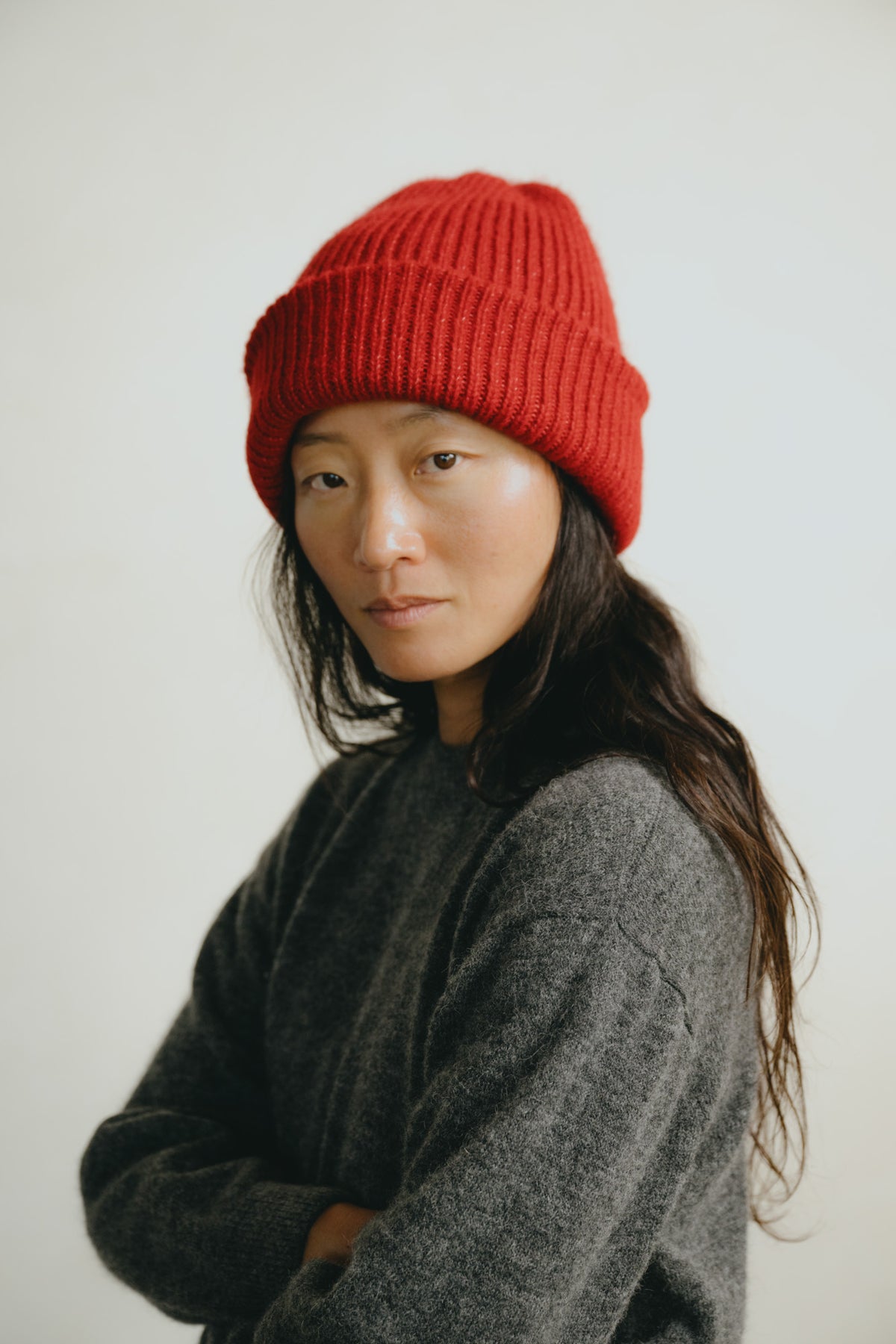 A woman wearing a bright red ribbed knit beanie and a dark gray knit sweater, photographed against a plain light background.