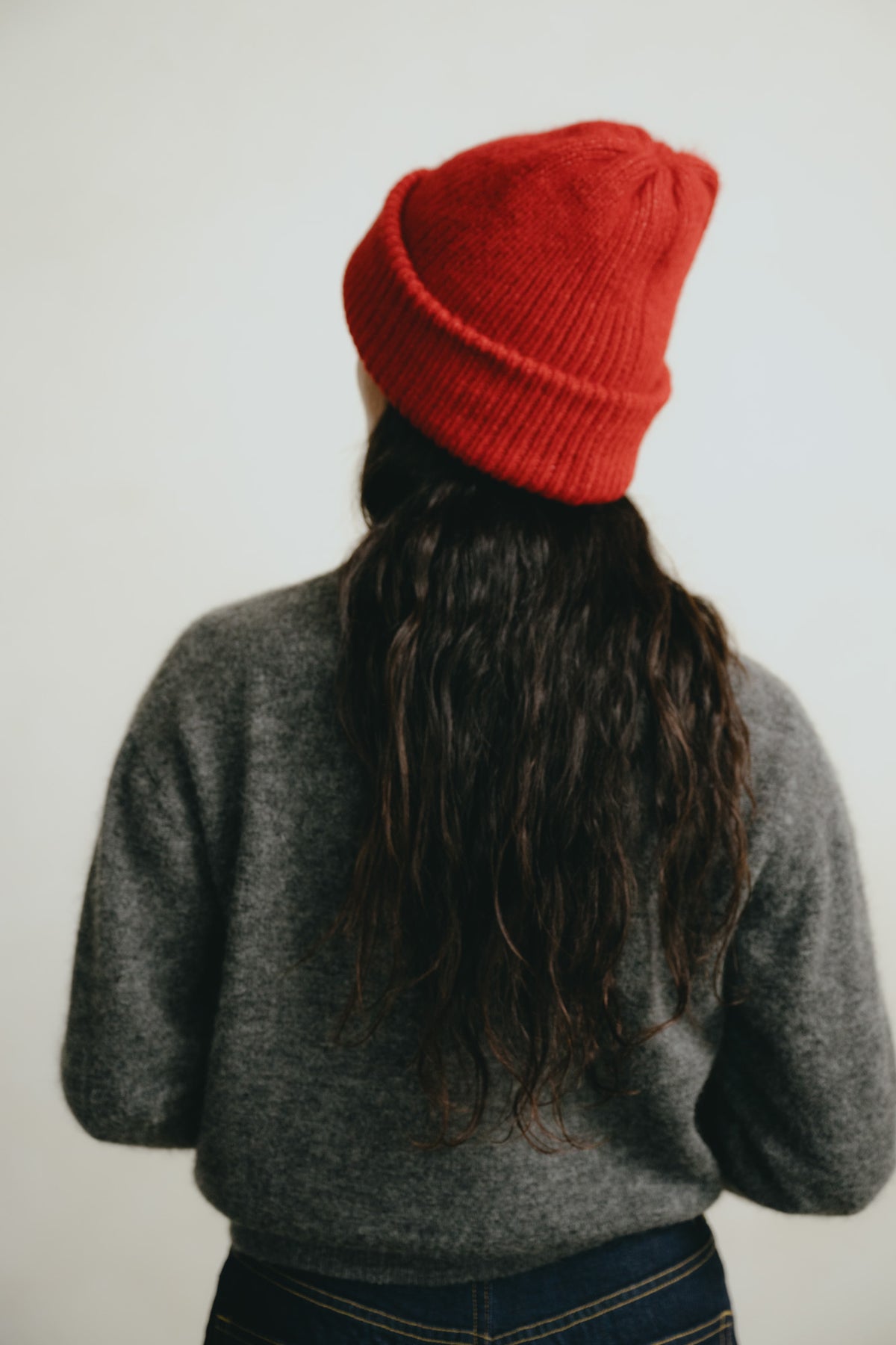 A person shown from behind wearing a bright red knit beanie, a gray knit sweater, and dark blue jeans against a plain light gray background.