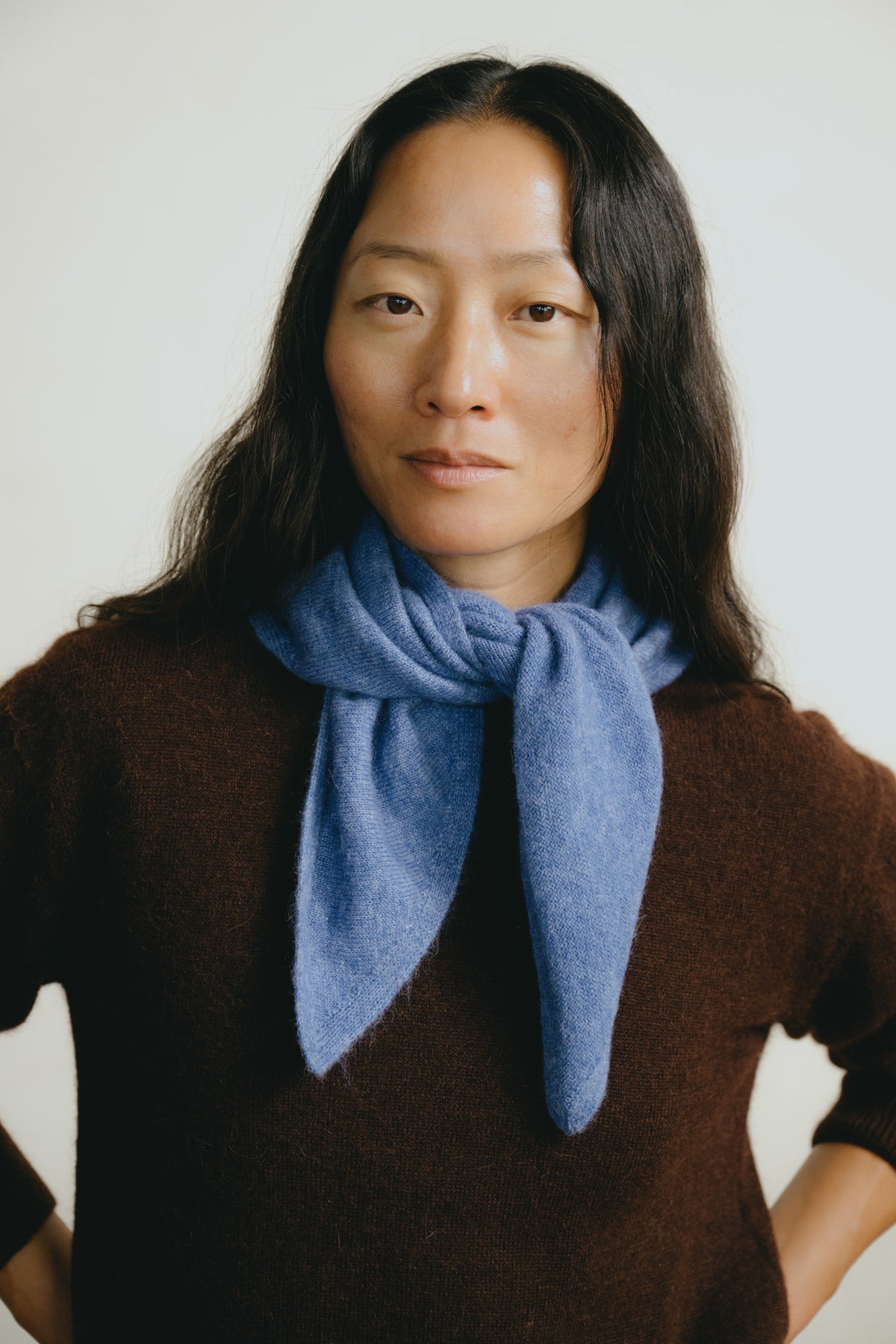 A woman with long dark hair wearing a brown knit sweater and a soft blue scarf, photographed against a plain light background.