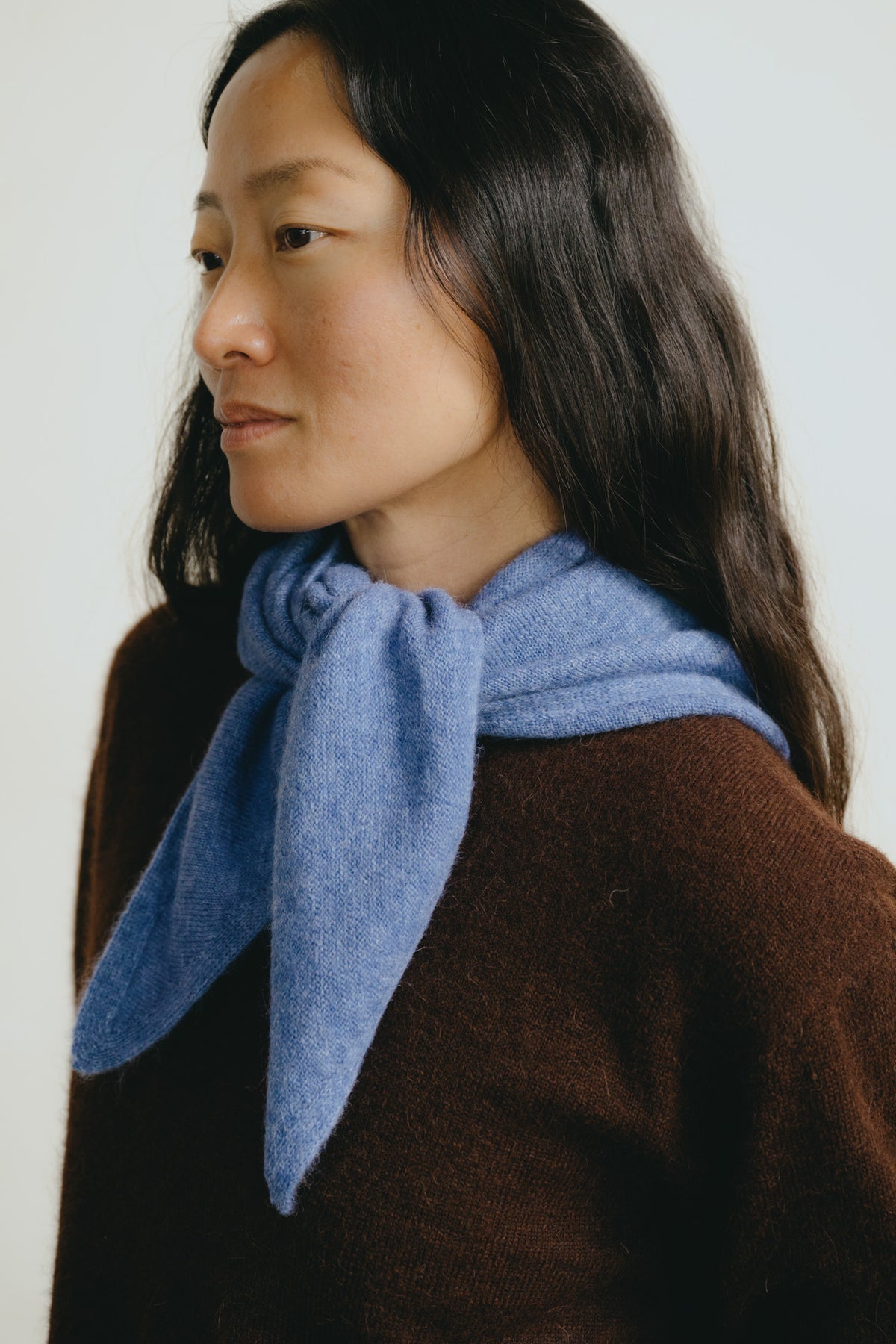 A woman with long dark hair is shown in profile wearing a soft, light blue knit scarf tied around her neck over a brown sweater, against a plain light background.