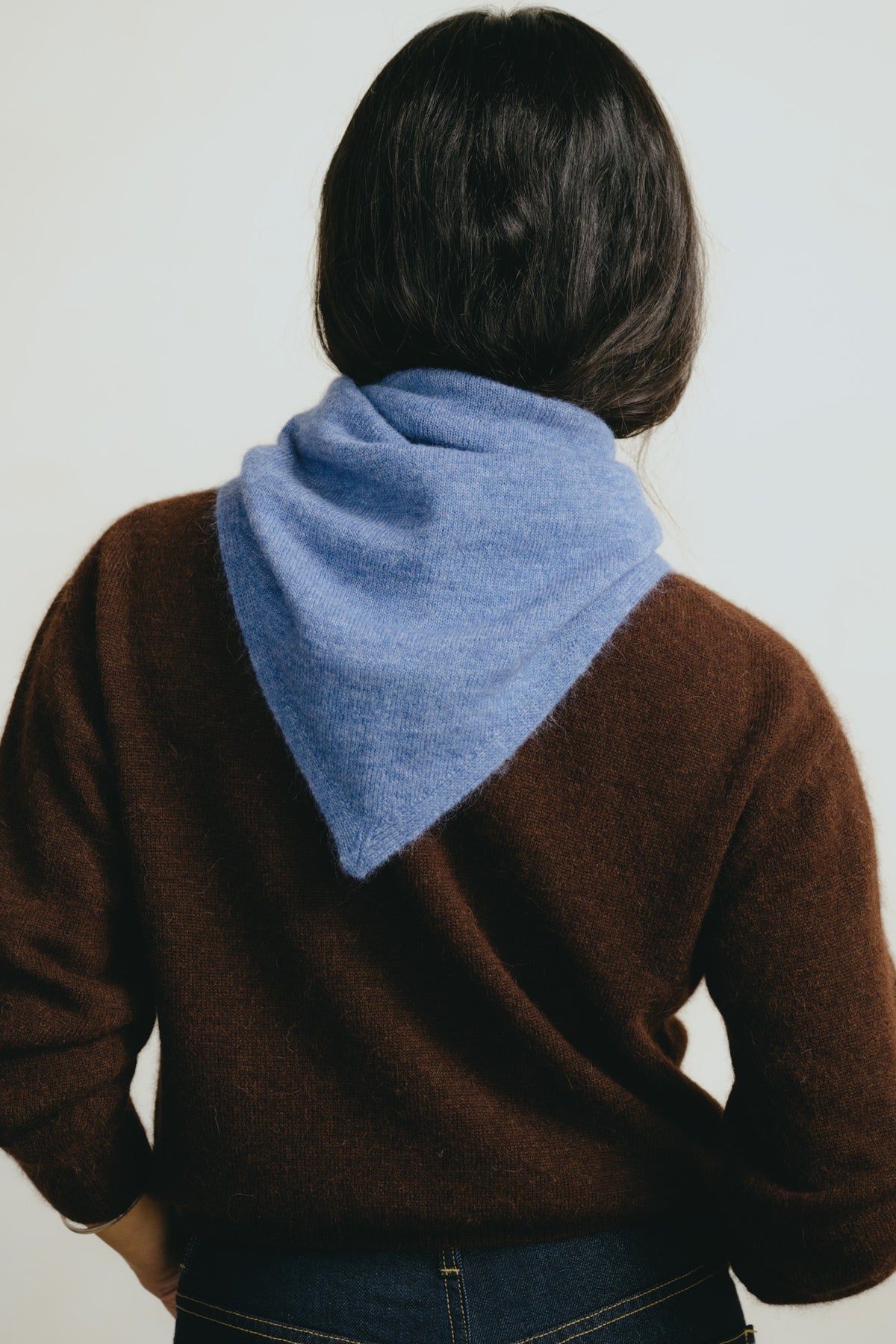 A person viewed from behind wearing a brown knit sweater and a light blue triangular neck warmer or bandana-style wrap around the neck, against a plain light background.