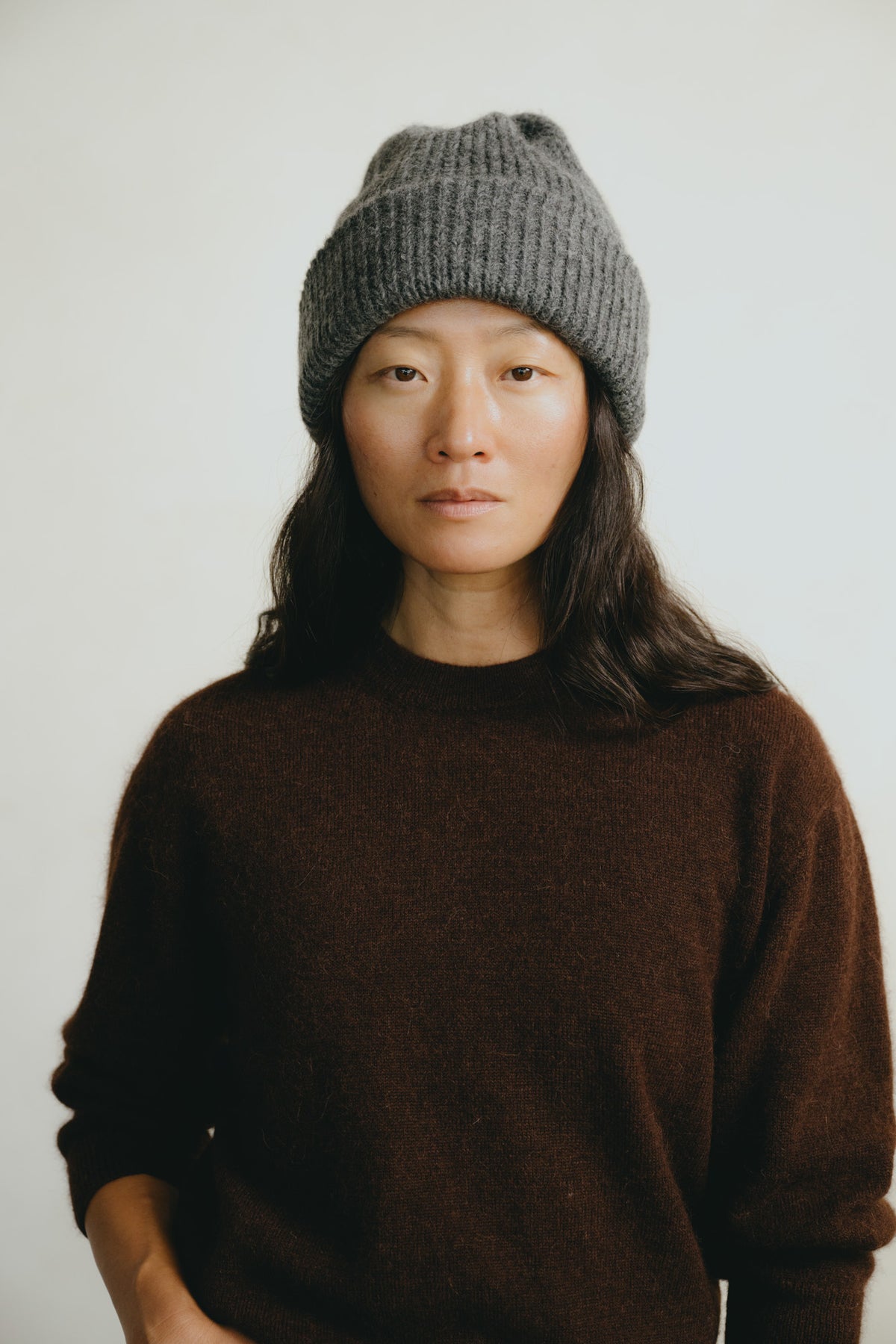 A woman wearing a dark brown knit crewneck sweater and a gray ribbed beanie hat, photographed against a plain light gray background.