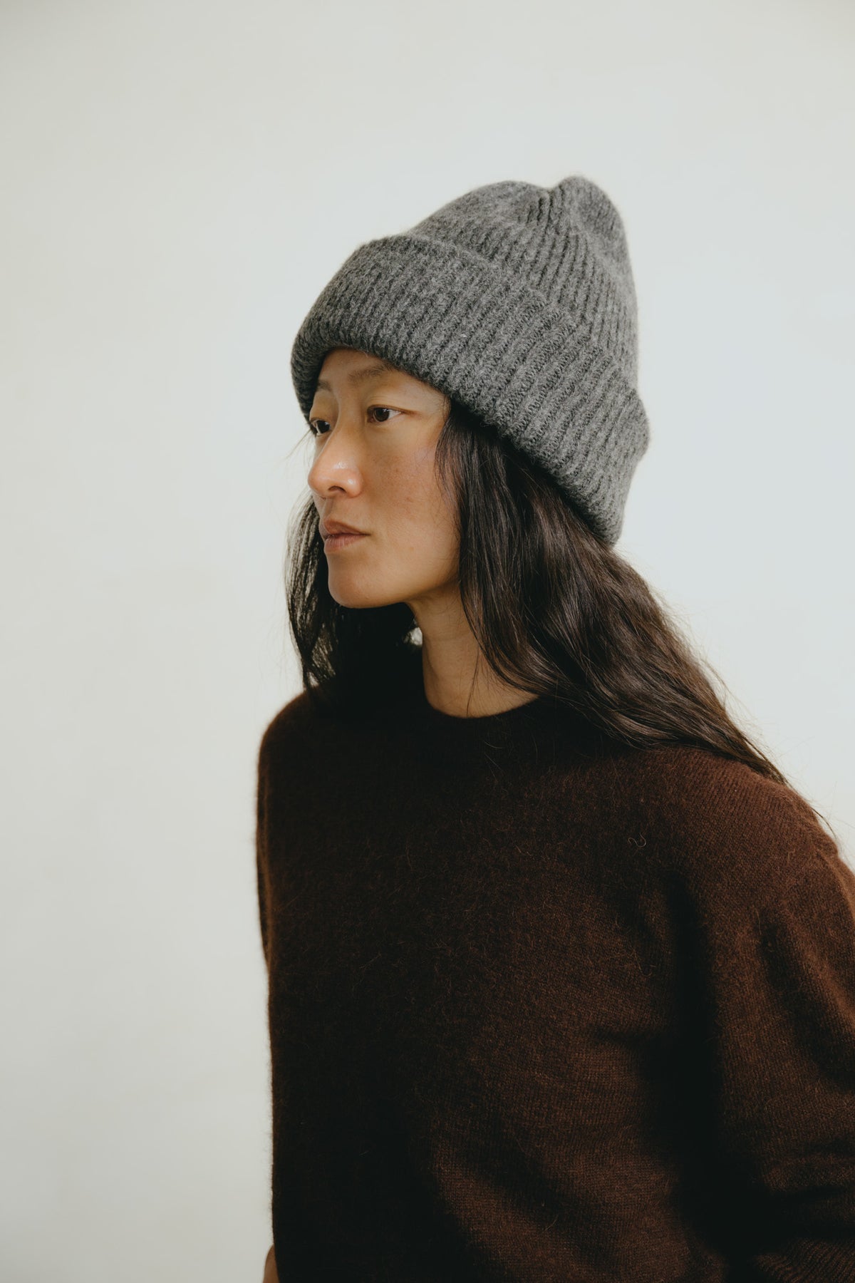 A woman in profile wearing a gray ribbed knit beanie and a dark brown sweater, photographed against a plain light background.