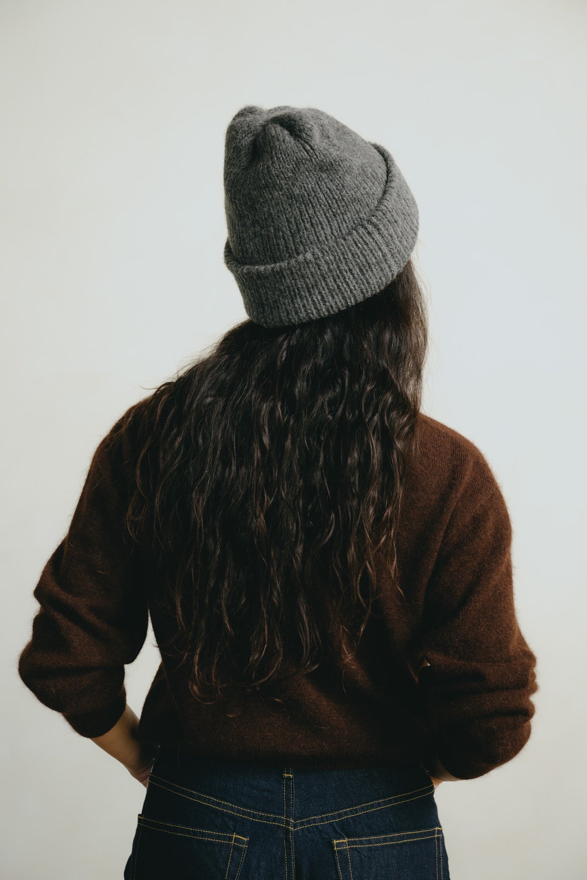 A person viewed from behind wearing a gray ribbed knit beanie, a brown sweater, and blue jeans against a plain light background.
