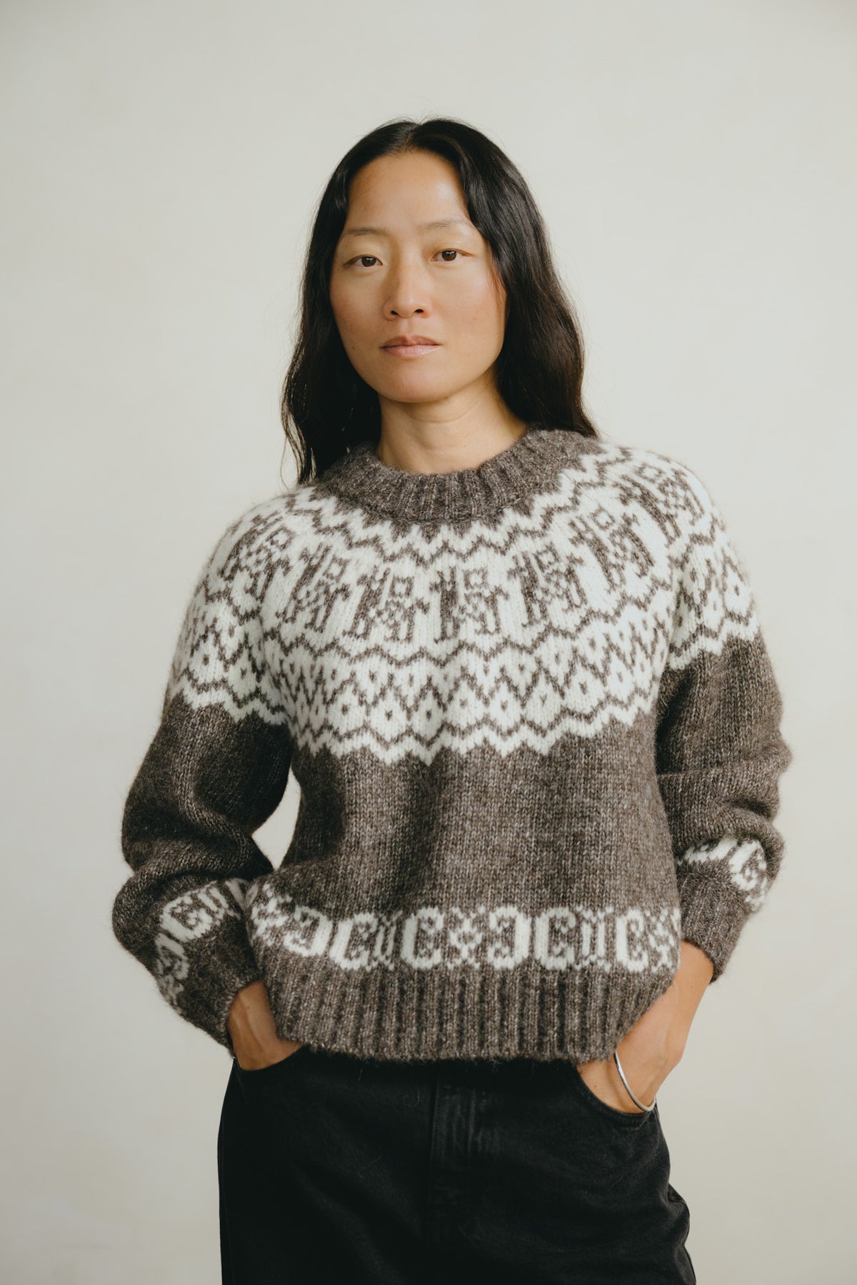 A woman wearing a brown and cream Fair Isle knit sweater with black pants, standing against a plain light gray background.