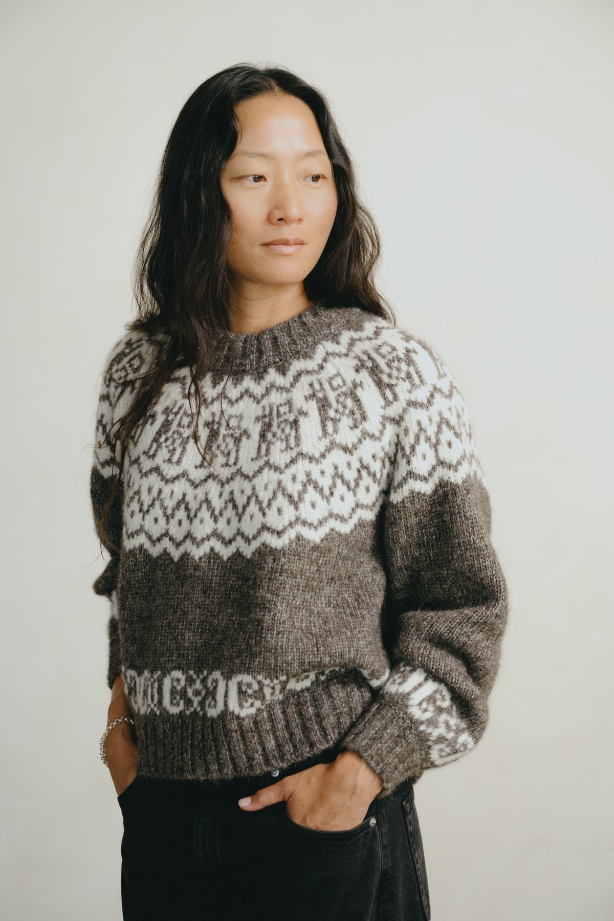 A woman wearing a brown and cream Fair Isle knit sweater with 'M.CXJC' text on the hem, standing against a plain light background.