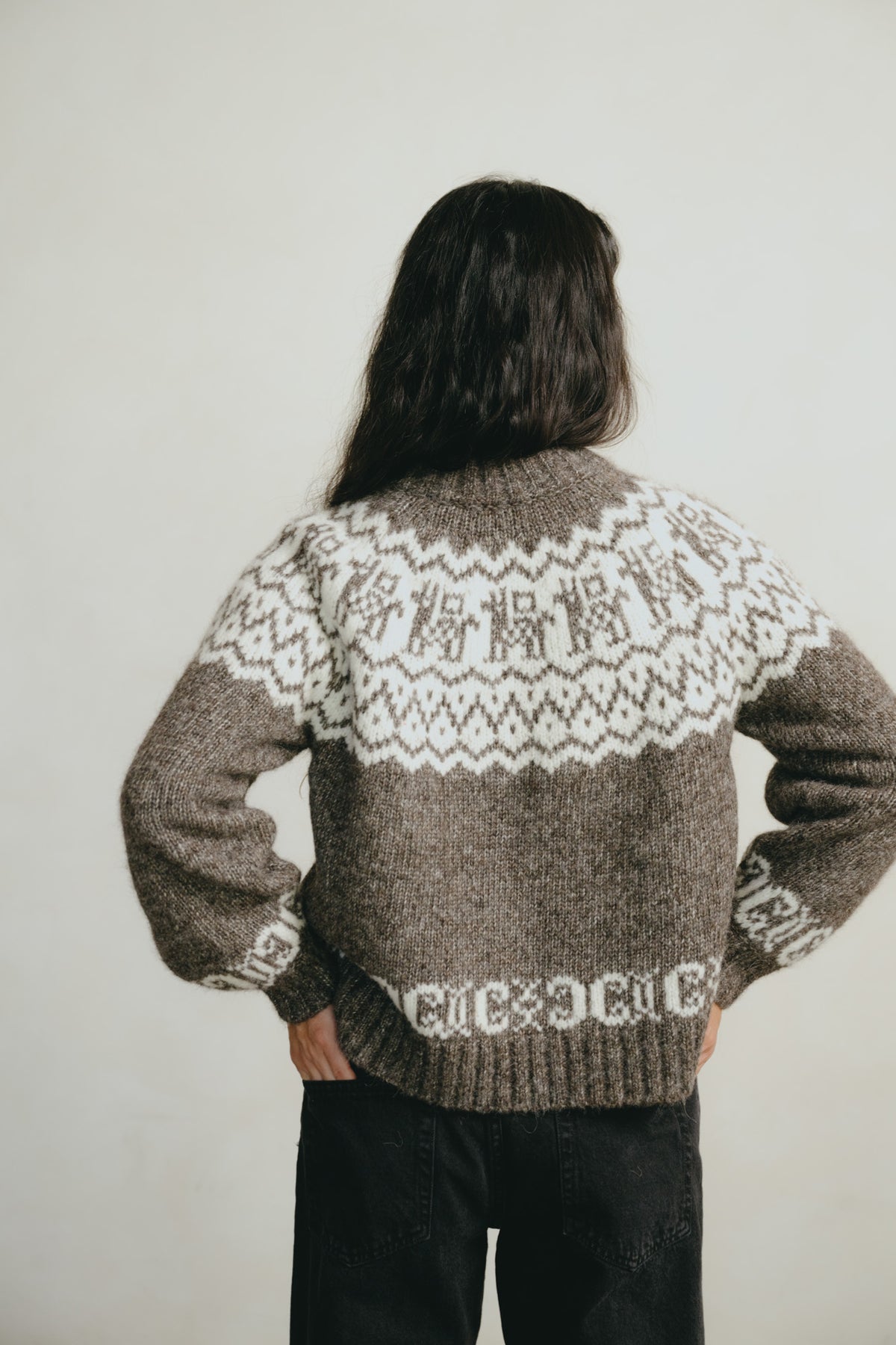 A person viewed from behind wearing a brown and cream Fair Isle knit sweater with Japanese characters knitted across the upper back, paired with black pants, against a plain light background.