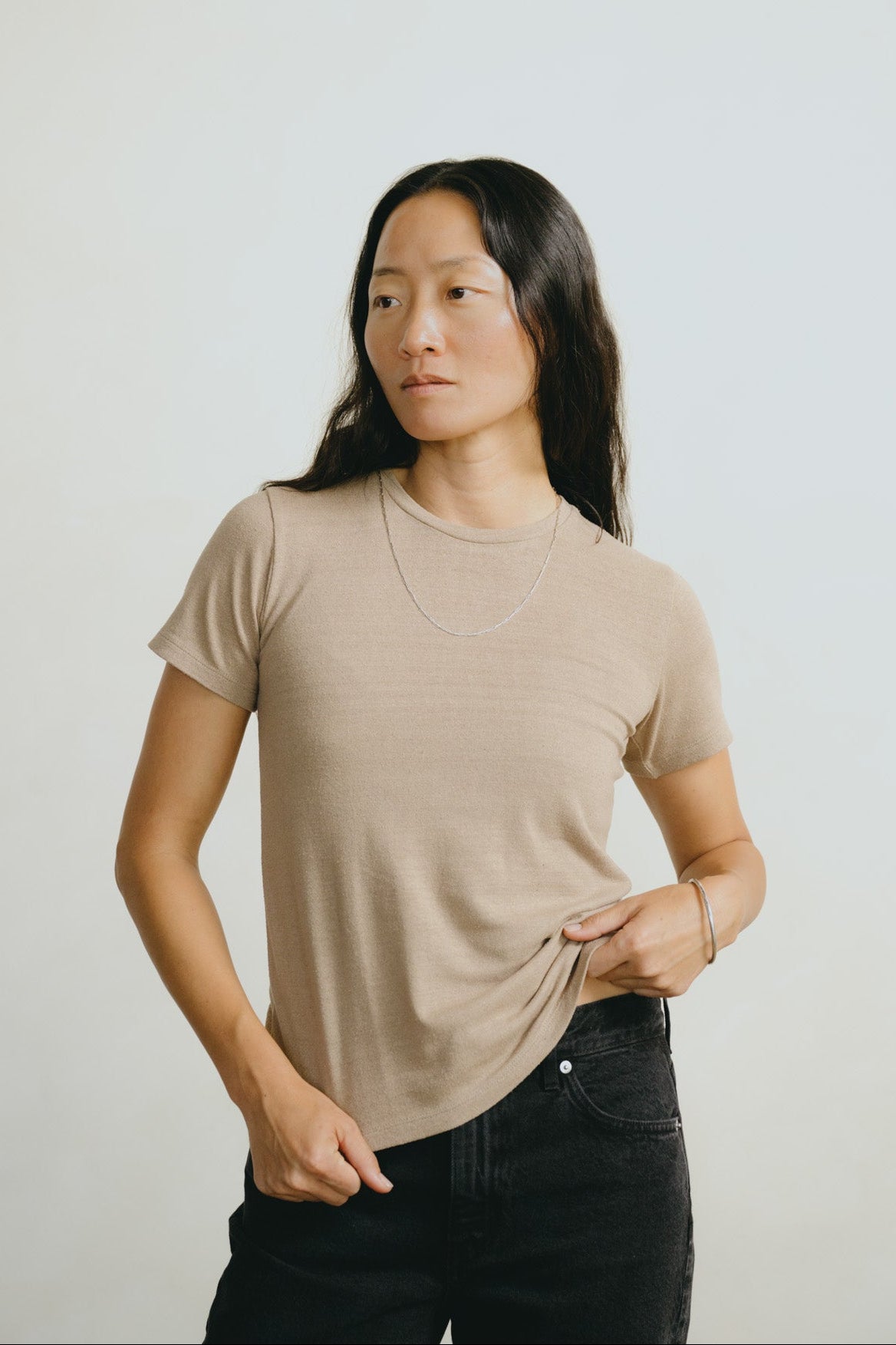 A woman modeling a light beige crewneck t-shirt paired with black jeans, standing against a plain light gray studio background.