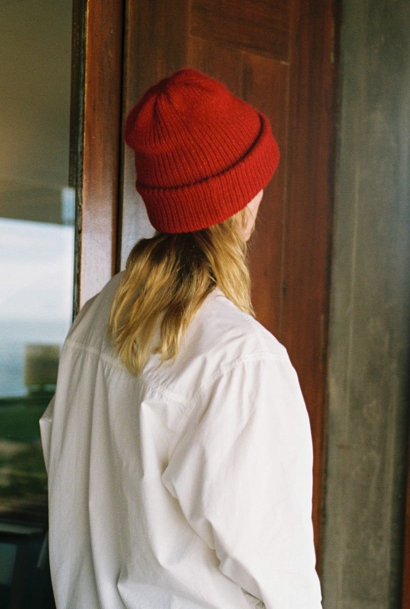 A person viewed from behind wearing a bright red ribbed knit beanie and an oversized white shirt, standing in front of a dark wooden door.