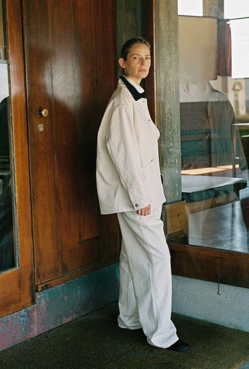 A model stands in profile wearing a white two-piece outfit consisting of a long-sleeve jacket with a black collar and matching wide-leg trousers, set against a rustic background with a wooden door and mirror.