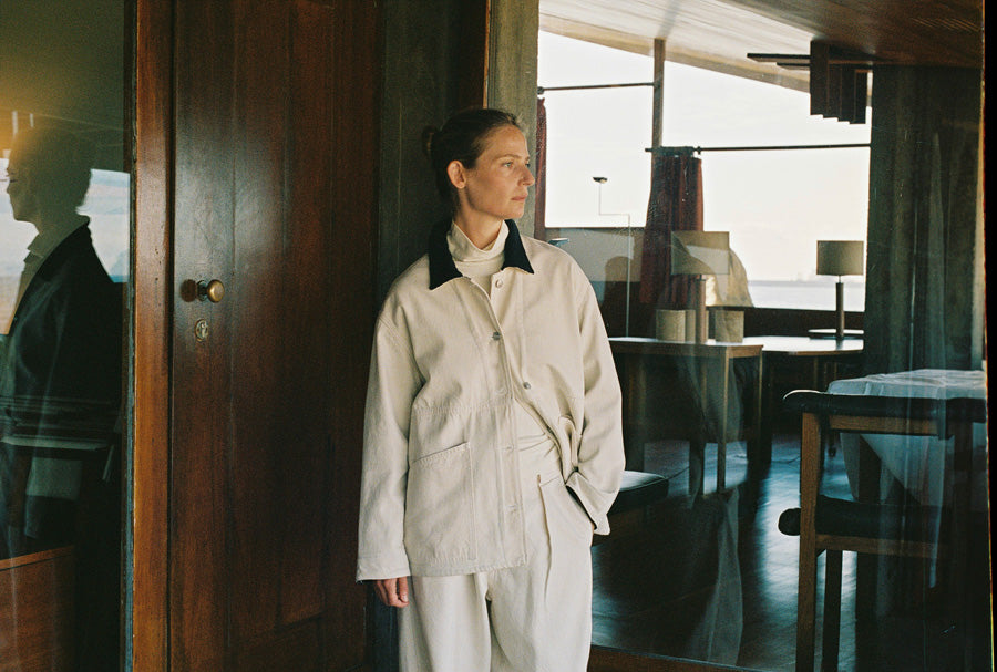 A woman stands indoors near a large window, wearing a light cream or off-white jacket and matching pants with a dark collar. The room has dark wood furniture and glass doors, with natural light coming from outside.