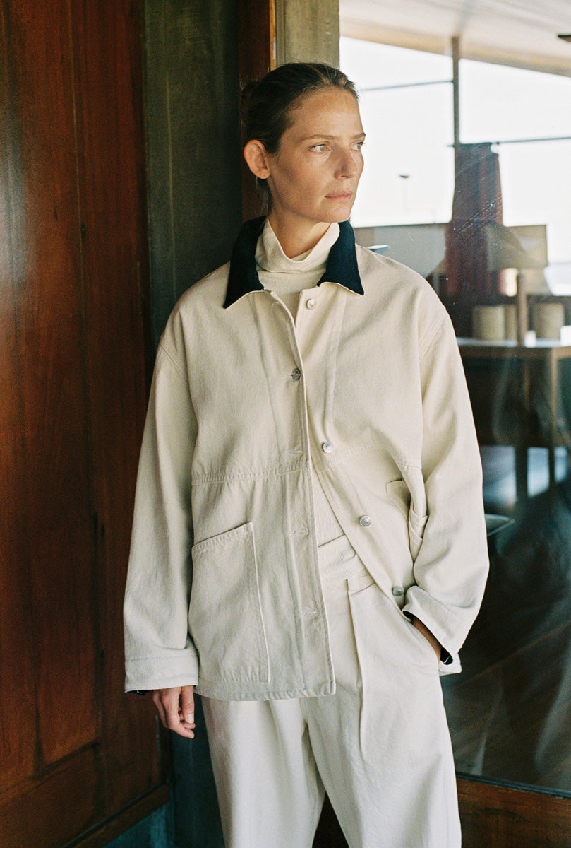 A woman wearing a light beige linen utility jacket with a dark navy collar, layered over a cream turtleneck and matching beige trousers, standing in a dimly lit room with a wooden door.