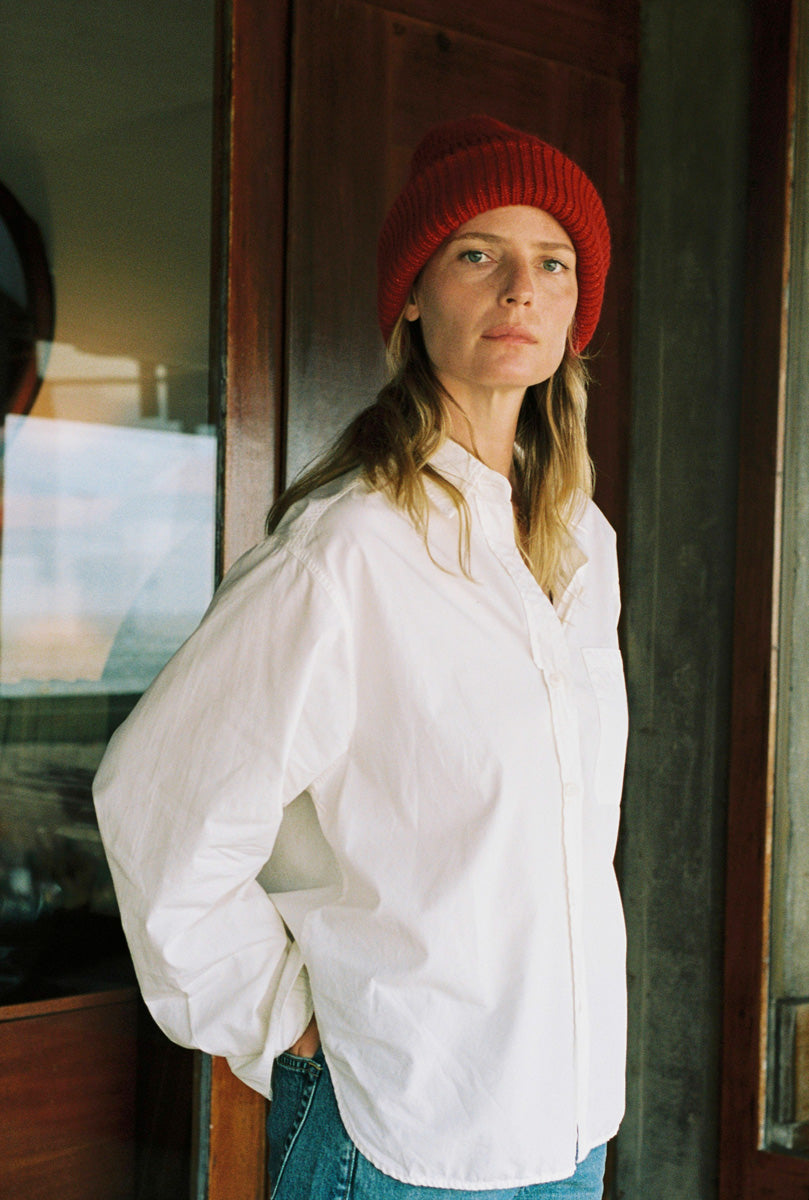 A woman wearing a red knit beanie and an oversized white button-up shirt, standing indoors near a dark wooden door.