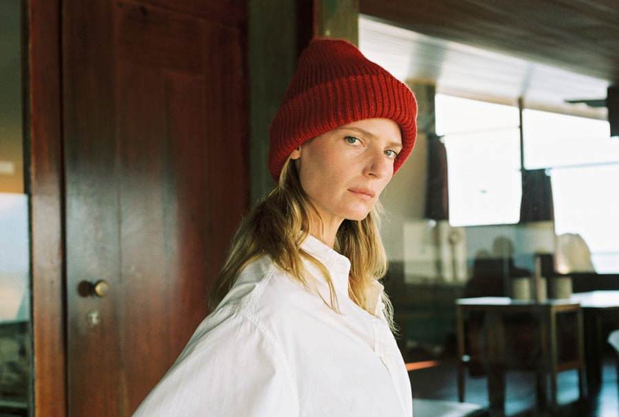 A woman wearing a red knit beanie and an oversized white button-up shirt, standing indoors near a dark wooden door.