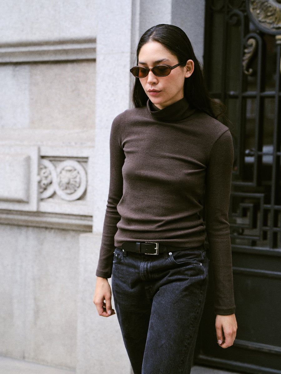 A woman standing on the street with sunglasses and wearing a long-sleeved dark brown turtleneck and black jeans.