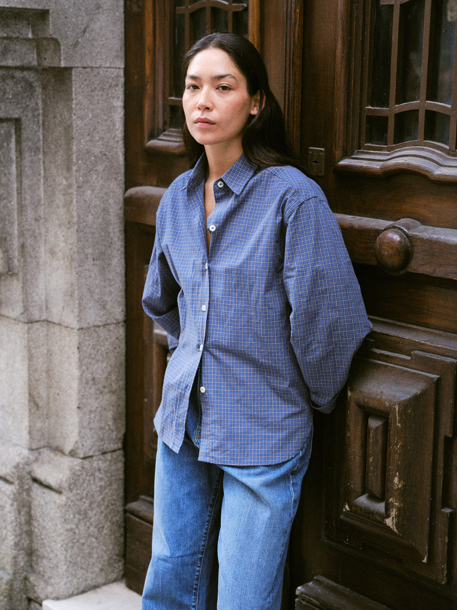 A woman wearing a blue and black checkered button-down shirt and blue jeans, standing in front of a dark wooden door and stone wall.