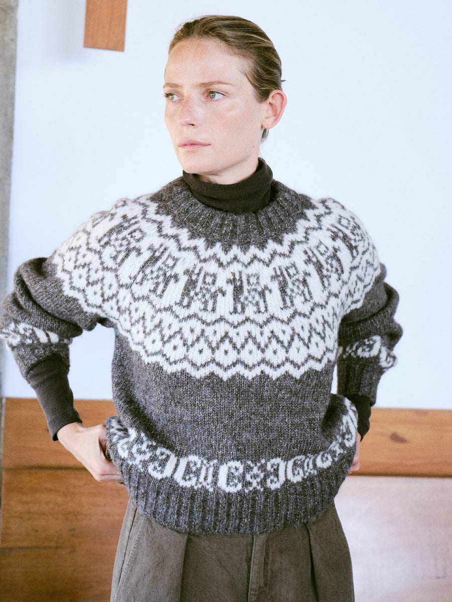 A woman wearing a dark gray and white Fair Isle knit sweater over a black turtleneck, standing indoors against a light wall with wooden paneling.
