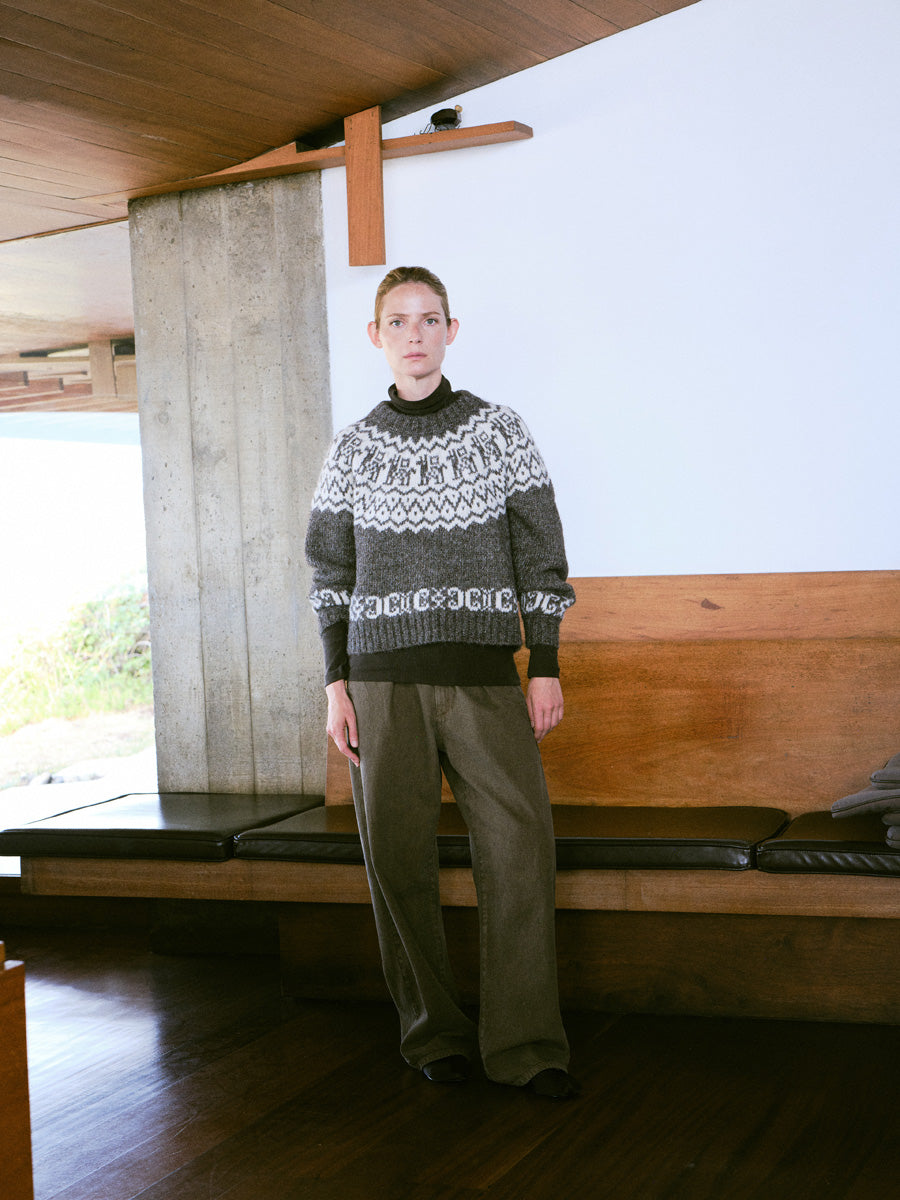 A model stands in a modern interior space wearing a dark gray and white patterned knit sweater with dark olive pants.