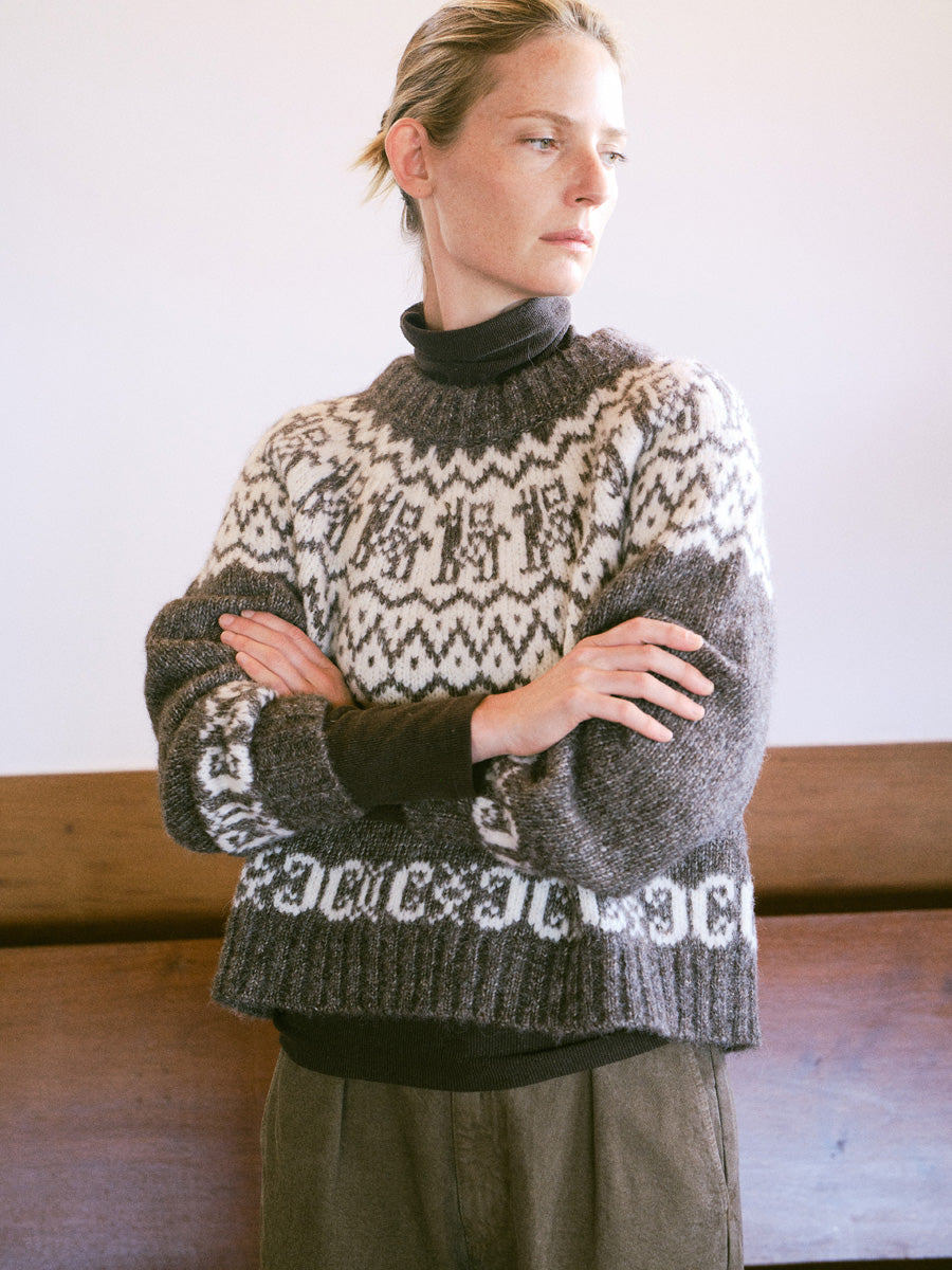 A woman wearing a dark gray and cream Fair Isle knit turtleneck sweater, standing with arms crossed in front of a wooden bench.