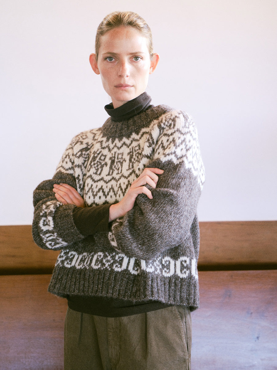A woman wearing a dark gray and cream Fair Isle knit sweater over a black turtleneck, standing with arms crossed in front of a plain light wall and wooden bench.
